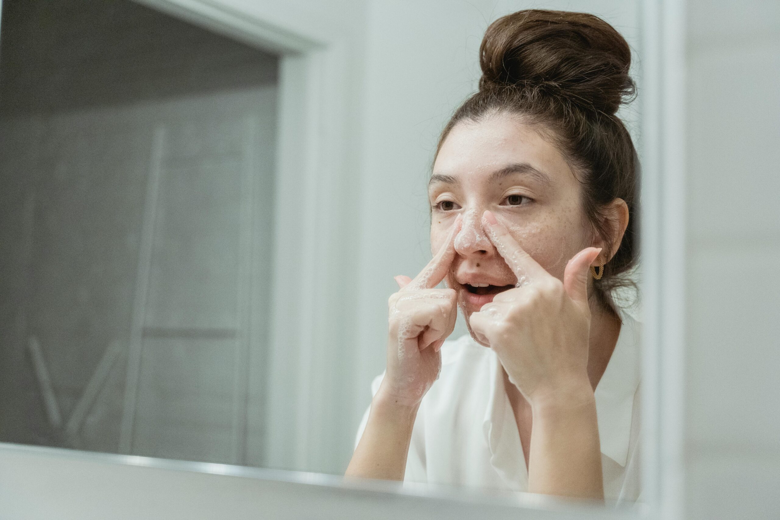 A young woman applies skincare in front of a bathroom mirror, focusing on facial care.